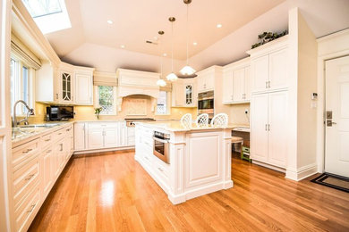Example of a classic medium tone wood floor eat-in kitchen design in New York with an undermount sink, raised-panel cabinets, white cabinets, granite countertops, beige backsplash, stone tile backsplash, paneled appliances and an island