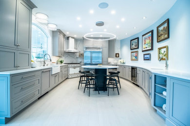 This is an example of a medium sized modern kitchen/diner in Portland Maine with a belfast sink, shaker cabinets, grey cabinets, engineered stone countertops, multi-coloured splashback, engineered quartz splashback, stainless steel appliances, light hardwood flooring, an island, white floors and multicoloured worktops.