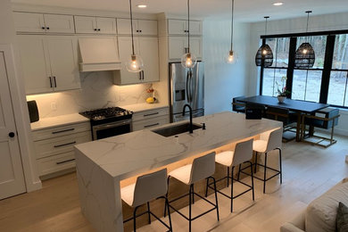 Photo of a small classic single-wall kitchen/diner in Portland Maine with a submerged sink, shaker cabinets, white cabinets, engineered stone countertops, white splashback, stone tiled splashback, stainless steel appliances, light hardwood flooring, an island, brown floors and white worktops.