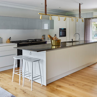 Inspiration for a contemporary galley open plan kitchen in Sussex with light hardwood flooring, an island, flat-panel cabinets, white cabinets, black appliances and brown floors.