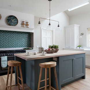 This is an example of a country l-shaped kitchen in Other with a farmhouse sink, recessed-panel cabinets, green cabinets, wood benchtops, green splashback, black appliances, medium hardwood floors, with island, beige floor and beige benchtop.