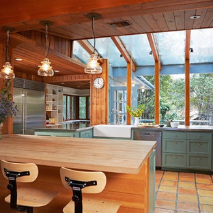 Photo of a country u-shaped kitchen in Los Angeles with terra-cotta floors, a farmhouse sink, shaker cabinets, green cabinets, stainless steel benchtops, stainless steel appliances, a peninsula and orange floor.
