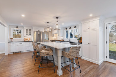 Photo of a large farmhouse l-shaped kitchen/diner in Boston with a belfast sink, shaker cabinets, white cabinets, engineered stone countertops, white splashback, metro tiled splashback, stainless steel appliances, medium hardwood flooring, an island, brown floors and white worktops.