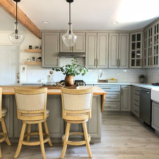 Photo of a small country l-shaped open plan kitchen in Los Angeles with raised-panel cabinets, grey cabinets, with island, a farmhouse sink, quartz benchtops, white splashback, subway tile splashback, stainless steel appliances, light hardwood floors, beige floor and white benchtop.