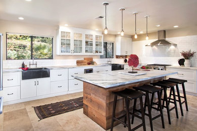 Example of a mid-sized transitional u-shaped travertine floor and beige floor open concept kitchen design in San Diego with a farmhouse sink, flat-panel cabinets, white cabinets, marble countertops, white backsplash, subway tile backsplash, stainless steel appliances and an island