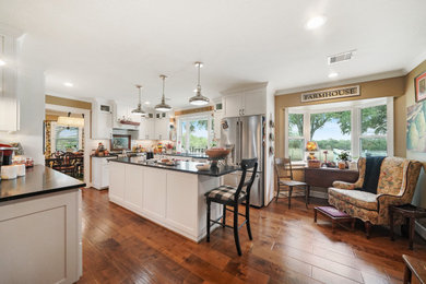 Example of a huge country u-shaped medium tone wood floor and brown floor open concept kitchen design in Austin with a farmhouse sink, shaker cabinets, white cabinets, granite countertops, white backsplash, ceramic backsplash, stainless steel appliances, an island and black countertops
