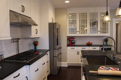 This is an example of a large classic u-shaped enclosed kitchen in DC Metro with a belfast sink, shaker cabinets, white cabinets, composite countertops, white splashback, metro tiled splashback, stainless steel appliances, dark hardwood flooring, an island, brown floors and black worktops.