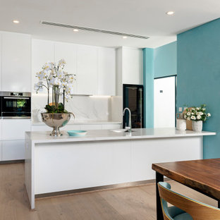 Inspiration for a mid-sized contemporary galley eat-in kitchen in Perth with an undermount sink, flat-panel cabinets, white cabinets, white splashback, black appliances, light hardwood floors, a peninsula, beige floor and white benchtop.
