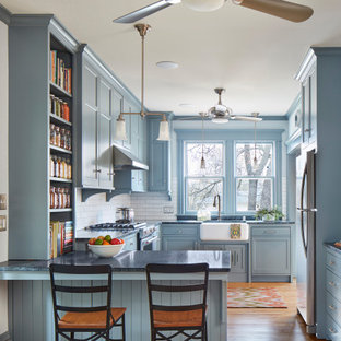 Diseño de cocina en U, de estilo americano, con fregadero sobremueble, armarios estilo shaker, puertas de armario azules, salpicadero blanco, salpicadero de azulejos tipo metro, electrodomésticos de acero inoxidable, suelo de madera clara, península y encimeras grises
