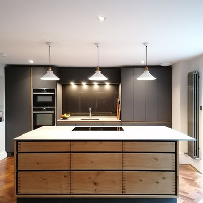 Example of a large trendy galley brown floor kitchen design in Hertfordshire with an undermount sink, flat-panel cabinets, gray cabinets, black backsplash, glass sheet backsplash, paneled appliances, an island and white countertops