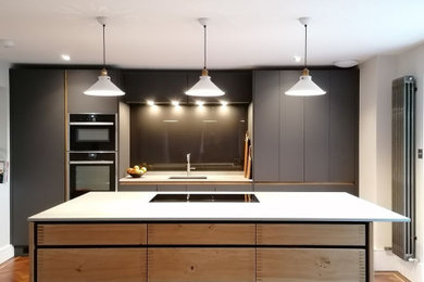 Inspiration for a large contemporary galley kitchen in Hertfordshire with a submerged sink, flat-panel cabinets, grey cabinets, black splashback, glass sheet splashback, integrated appliances, an island, brown floors and white worktops.