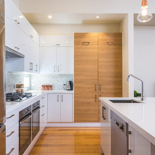 This is an example of a mid-sized contemporary u-shaped open plan kitchen in Melbourne with an undermount sink, solid surface benchtops, white splashback, stone tile splashback, stainless steel appliances, medium hardwood floors, with island, white benchtop, flat-panel cabinets, white cabinets and brown floor.