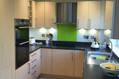 Design ideas for a small modern u-shaped enclosed kitchen in Hampshire with an integrated sink, flat-panel cabinets, light wood cabinets, green splashback, glass sheet splashback, stainless steel appliances, ceramic flooring, beige floors and black worktops.