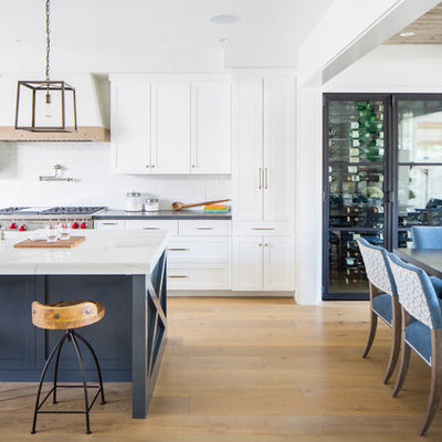 Example of a large cottage galley medium tone wood floor and brown floor open concept kitchen design in Orange County with a farmhouse sink, shaker cabinets, white cabinets, white backsplash, subway tile backsplash, stainless steel appliances, an island and white countertops