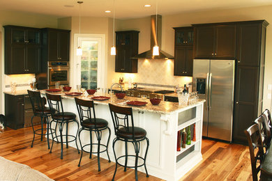 ミルウォーキーにあるトラディショナルスタイルのおしゃれなアイランドキッチン (落し込みパネル扉のキャビネット、ベージュキッチンパネル、シルバーの調理設備、淡色無垢フローリング) の写真