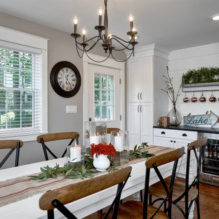 Photo of a mid-sized country single-wall open plan kitchen in Chicago with a farmhouse sink, shaker cabinets, white cabinets, granite benchtops, white splashback, stainless steel appliances, medium hardwood floors, with island, brown floor, black benchtop and shiplap splashback.