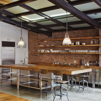 Inspiration for a mid-sized industrial l-shaped concrete floor and gray floor eat-in kitchen remodel in New York with stainless steel appliances, stainless steel cabinets, wood countertops, flat-panel cabinets, red backsplash, brick backsplash, an island and a farmhouse sink