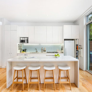 This is an example of a transitional single-wall kitchen in Sydney with an undermount sink, flat-panel cabinets, white cabinets, granite benchtops, green splashback, glass sheet splashback, stainless steel appliances, medium hardwood floors, with island, brown floor and white benchtop.