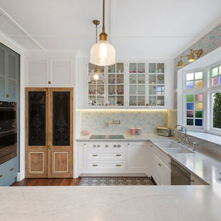 Photo of a transitional u-shaped kitchen in Melbourne with a farmhouse sink, louvered cabinets, white cabinets, multi-coloured splashback, mosaic tile splashback, stainless steel appliances, dark hardwood floors, a peninsula, brown floor and grey benchtop.