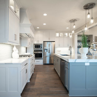 Photo of an arts and crafts l-shaped open plan kitchen in Kansas City with a drop-in sink, white cabinets, white splashback, stainless steel appliances, medium hardwood floors, with island, brown floor and white benchtop.