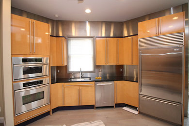Modelo de cocina moderna con armarios con paneles lisos, puertas de armario de madera clara, encimera de granito, salpicadero metalizado, salpicadero de metal, electrodomésticos de acero inoxidable y encimeras negras