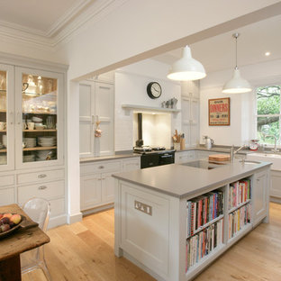 Photo of a medium sized traditional l-shaped kitchen/diner in Other with a belfast sink, recessed-panel cabinets, grey cabinets, white splashback, metro tiled splashback, light hardwood flooring, an island, beige floors, grey worktops and black appliances.