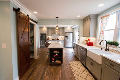 Large transitional l-shaped medium tone wood floor and brown floor enclosed kitchen photo in Cincinnati with a farmhouse sink, recessed-panel cabinets, gray cabinets, marble countertops, white backsplash, porcelain backsplash, stainless steel appliances and an island