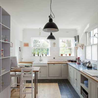 Example of a mid-sized classic l-shaped black floor eat-in kitchen design in London with shaker cabinets, gray cabinets, wood countertops, white backsplash, subway tile backsplash, brown countertops and a farmhouse sink