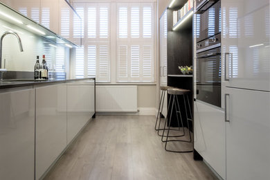 Inspiration for a small galley enclosed kitchen in London with a single-bowl sink, white cabinets, quartz worktops, grey splashback, glass sheet splashback, black appliances, laminate floors, a breakfast bar and grey floors.