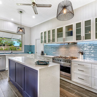 This is an example of a beach style l-shaped kitchen in Brisbane with an undermount sink, glass-front cabinets, white cabinets, blue splashback, subway tile splashback, stainless steel appliances, with island, brown floor, grey benchtop and timber.