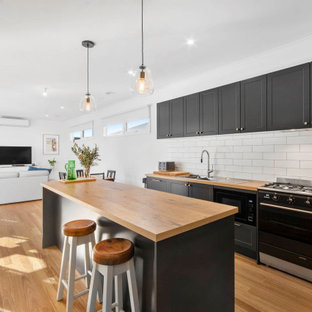 Inspiration for a transitional kitchen in Geelong with a double-bowl sink, wood benchtops, white splashback, ceramic splashback, black appliances, medium hardwood floors, brown benchtop, shaker cabinets, black cabinets, with island and brown floor.
