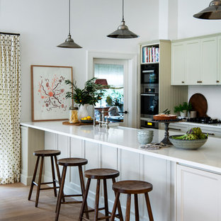 Design ideas for a traditional galley kitchen in Sydney with a farmhouse sink, recessed-panel cabinets, white cabinets, white splashback, black appliances, medium hardwood floors, a peninsula, brown floor and white benchtop.