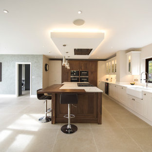 Design ideas for a classic kitchen/diner in Berkshire with a belfast sink, glass-front cabinets, beige cabinets, black appliances, an island, beige floors and white worktops.