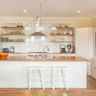 Photo of a country l-shaped kitchen in Other with a drop-in sink, flat-panel cabinets, white cabinets, wood benchtops, white splashback, subway tile splashback, stainless steel appliances, medium hardwood floors, with island, brown floor and brown benchtop.