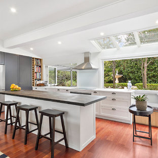 This is an example of an expansive contemporary l-shaped open plan kitchen in Sydney with brown floor, shaker cabinets, with island, an integrated sink, white cabinets, zinc benchtops, white splashback, glass sheet splashback, black appliances, light hardwood floors and grey benchtop.