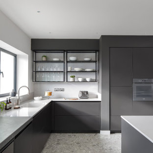 This is an example of an expansive contemporary grey and white l-shaped open plan kitchen in Sussex with flat-panel cabinets, black cabinets, an island, a single-bowl sink, integrated appliances, grey floors and white worktops.