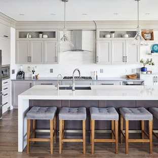 Imagen de cocina tradicional renovada, grande, con armarios estilo shaker, puertas de armario grises, salpicadero blanco, electrodomésticos de acero inoxidable, una isla, suelo marrón, fregadero bajoencimera, suelo de madera oscura y encimeras grises