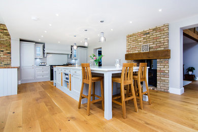 Photo of a large rural u-shaped kitchen in Hertfordshire with shaker cabinets, grey cabinets, quartz worktops, an island, a belfast sink, white splashback, integrated appliances, light hardwood flooring and white worktops.