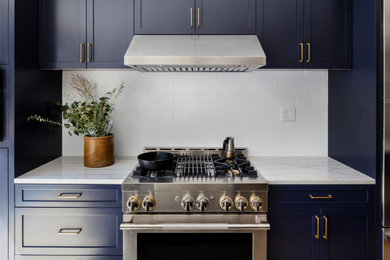 Bild på ett vit linjärt vitt kök, med en rustik diskho, skåp i shakerstil, blå skåp, marmorbänkskiva, vitt stänkskydd, stänkskydd i tunnelbanekakel, rostfria vitvaror, ljust trägolv, en köksö och beiget golv