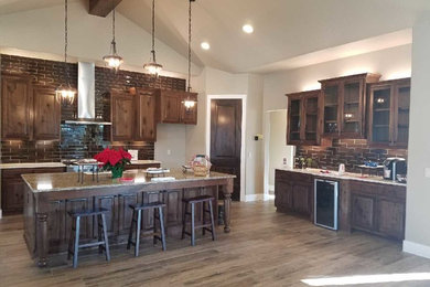 Large transitional l-shaped medium tone wood floor and brown floor kitchen photo in Austin with stainless steel appliances, a farmhouse sink, raised-panel cabinets, dark wood cabinets, granite countertops, brown backsplash, glass tile backsplash and an island