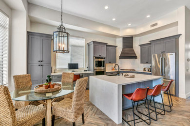 Large transitional l-shaped medium tone wood floor and brown floor eat-in kitchen photo in Phoenix with a farmhouse sink, shaker cabinets, gray cabinets, an island, white backsplash, stainless steel appliances and gray countertops