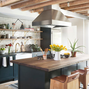 Inspiration for a large transitional galley open plan kitchen in New York with a farmhouse sink, open cabinets, white splashback, subway tile splashback, stainless steel appliances, light hardwood floors, with island, black cabinets, wood benchtops and beige floor.