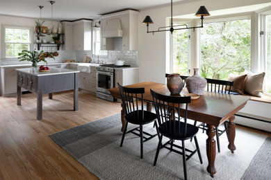 Example of a mid-sized cottage l-shaped light wood floor and beige floor eat-in kitchen design in Baltimore with a farmhouse sink, shaker cabinets, beige cabinets, quartz countertops, white backsplash, ceramic backsplash, stainless steel appliances, an island and white countertops