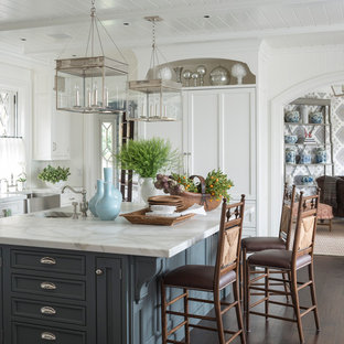 Example of a large classic u-shaped dark wood floor and brown floor kitchen design in Other with a farmhouse sink, recessed-panel cabinets, white cabinets, marble countertops, white backsplash, paneled appliances and an island