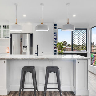This is an example of a large transitional u-shaped eat-in kitchen in Sunshine Coast with an undermount sink, shaker cabinets, white cabinets, quartzite benchtops, white splashback, ceramic splashback, stainless steel appliances, vinyl floors, with island, white benchtop and brown floor.