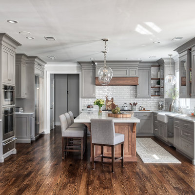 Inspiration for a transitional u-shaped medium tone wood floor and brown floor kitchen remodel in New York with a farmhouse sink, shaker cabinets, gray cabinets, white backsplash, subway tile backsplash, stainless steel appliances, an island and white countertops