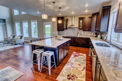 Open concept kitchen - mid-sized transitional l-shaped medium tone wood floor and brown floor open concept kitchen idea in Other with a single-bowl sink, shaker cabinets, dark wood cabinets, granite countertops, gray backsplash, porcelain backsplash, stainless steel appliances and an island