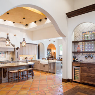 This is an example of a mediterranean u-shaped eat-in kitchen in Austin with a farmhouse sink, recessed-panel cabinets, light wood cabinets, multi-coloured splashback, mosaic tile splashback, stainless steel appliances, terra-cotta floors, with island, orange floor and beige benchtop.