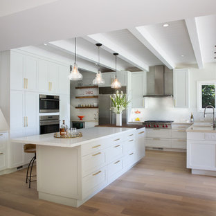 Diseño de cocina clásica renovada, grande, con armarios estilo shaker, puertas de armario blancas, salpicadero blanco, electrodomésticos de acero inoxidable, suelo de madera clara, una isla, suelo beige y encimeras blancas