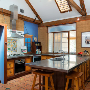 Mediterranean open plan kitchen in Brisbane with an integrated sink, flat-panel cabinets, stainless steel benchtops, multi-coloured splashback, stainless steel appliances, terra-cotta floors, a peninsula, red floor and grey benchtop.